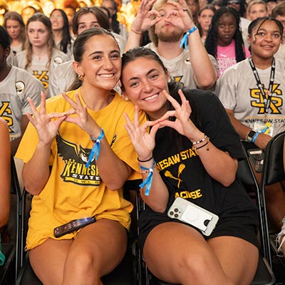 Two smiling women seated at an event, wearing Kennesaw State University shirts. They gesture owl eyes with their hands and are surrounded by a lively crowd.