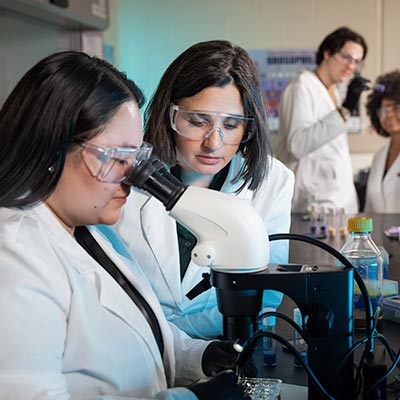 Two scientists in lab coats and goggles examine a sample under a microscope in a laboratory.